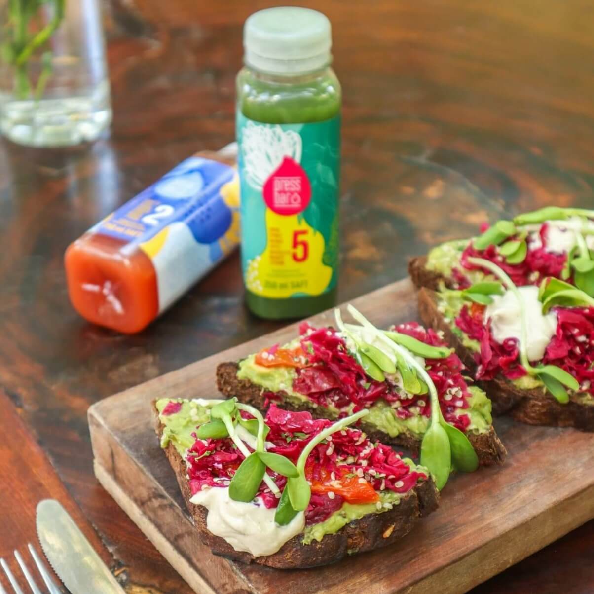 Vier scheiben Brot belegt mit Avocado  rotem und grünem Salat. Im hintergrund stehen zwei Flaschen voll mit Saft von pressbar. 